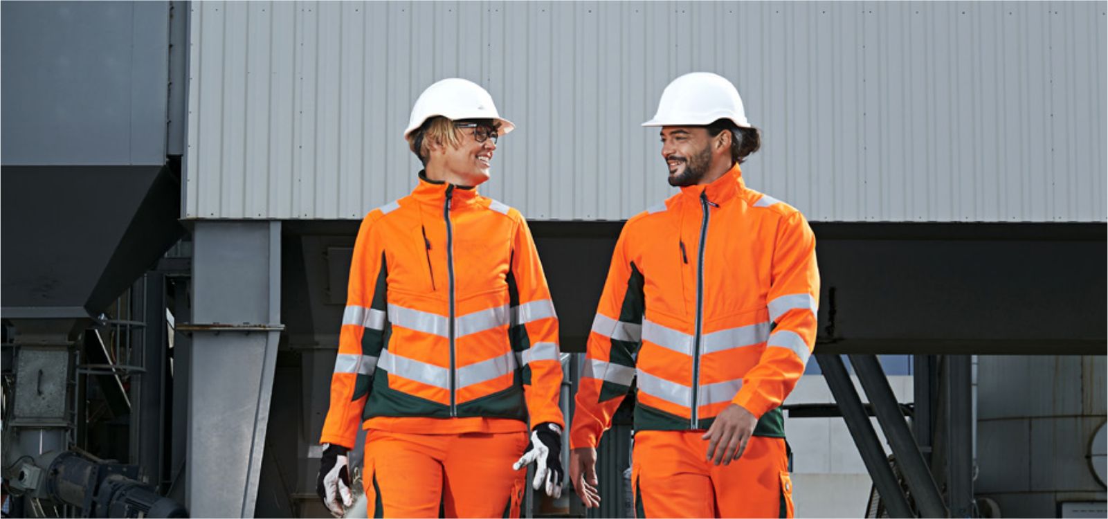 Männliche und weibliche Person, die auf die Kamera frontal zulaufen und orange-smaragd-farbene Warnschutzkleidung und weiße Helme tragen