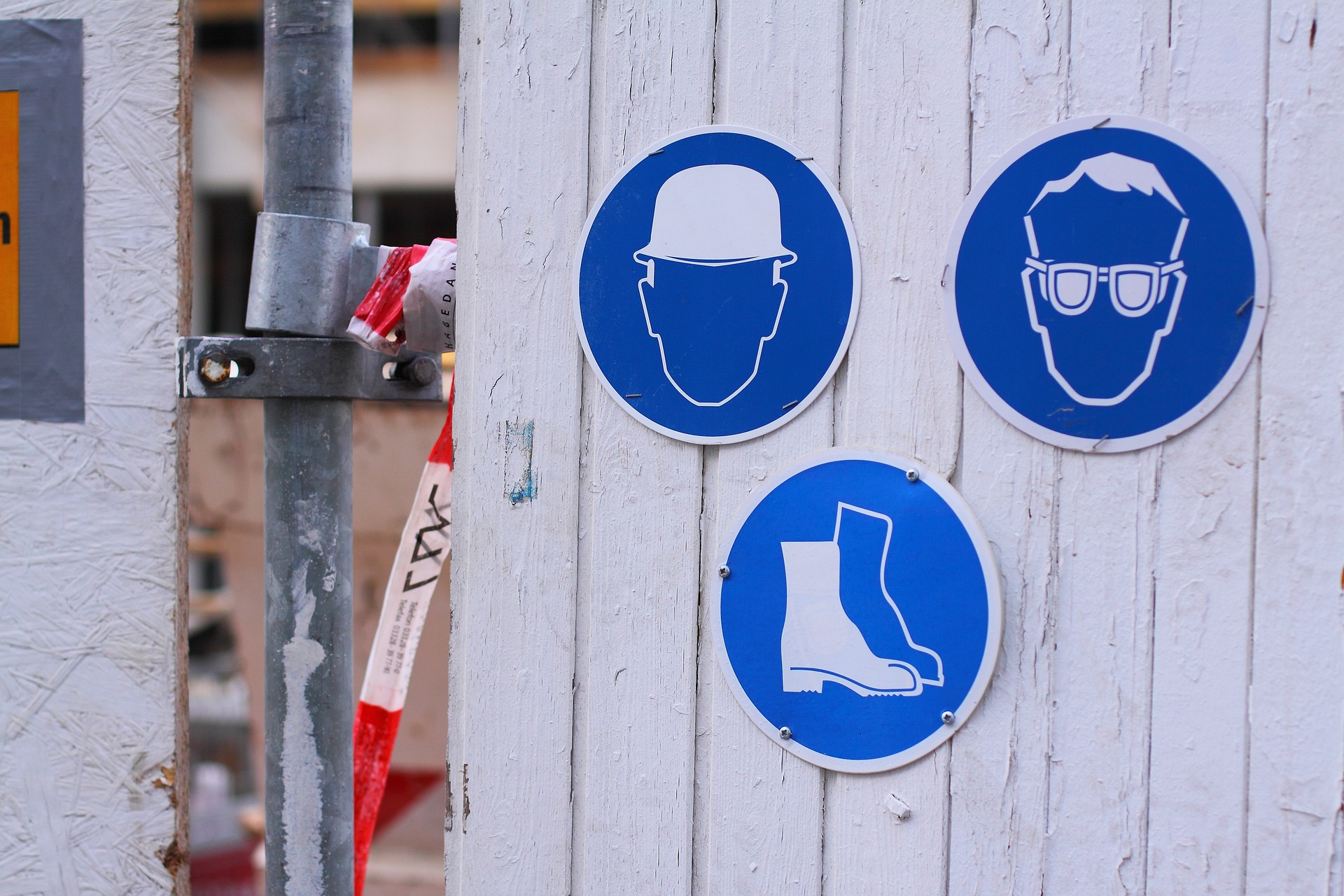 Drei Plaketten mit Symbolen an einer Holzwand hängend. Nr. 1: Ein Kopf mit weißem Schutzhelm; Nr. 2: Kopf mit Schutzbrille; Nr. 3: weiße Sicherheitsstiefel