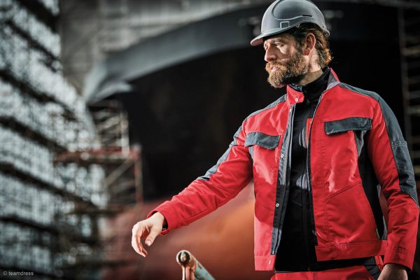 Männliche Person auf Baustelle, die rot-schwarze Arbeitskleidung sowie einen grauen Helm trägt
