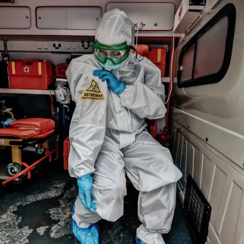 Frau in Krankenwagen, die Corona-Schutzanzug, Schutzbrille, Mundschutz und Handschuhe trägt